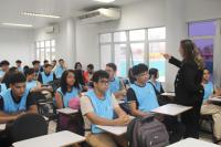 Imagem de uma sala de aula, com alunos sentados nas cadeiras, vestidos com colete azul. À frente, uma mulher, em pé, vestida com blaser e calça pretas, apontando para frente com a mão direita.